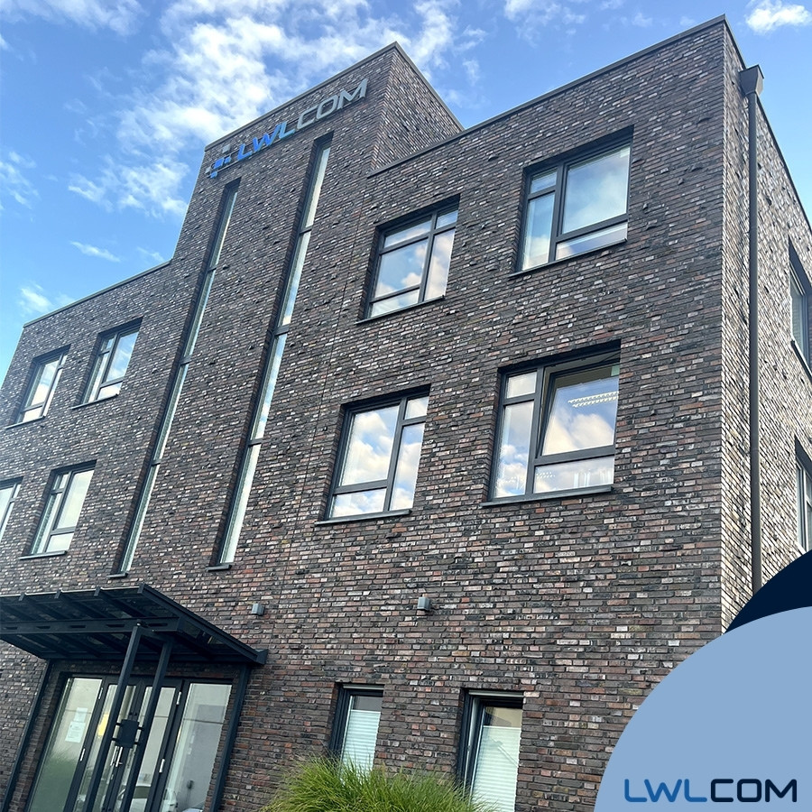 Seitenansicht des mehrstöckigen Firmengebäudes von LWLcom aus dunkelroten Ziegeln mit mehreren Fenstern und dem Firmenlogo unten rechts auf hellblauem Hintergrund.