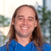 Ein lachender Mann mit langen braunen Haaren und einem blauem Kaputzenpullover.