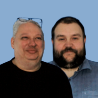 Zwei Männer, einer mit einem schwarzen Oberteil, Brille auf dem Kopf und grauen Haaren, der zweite mit dunklen Haaren und Vollbart. blaugraues Hemd.